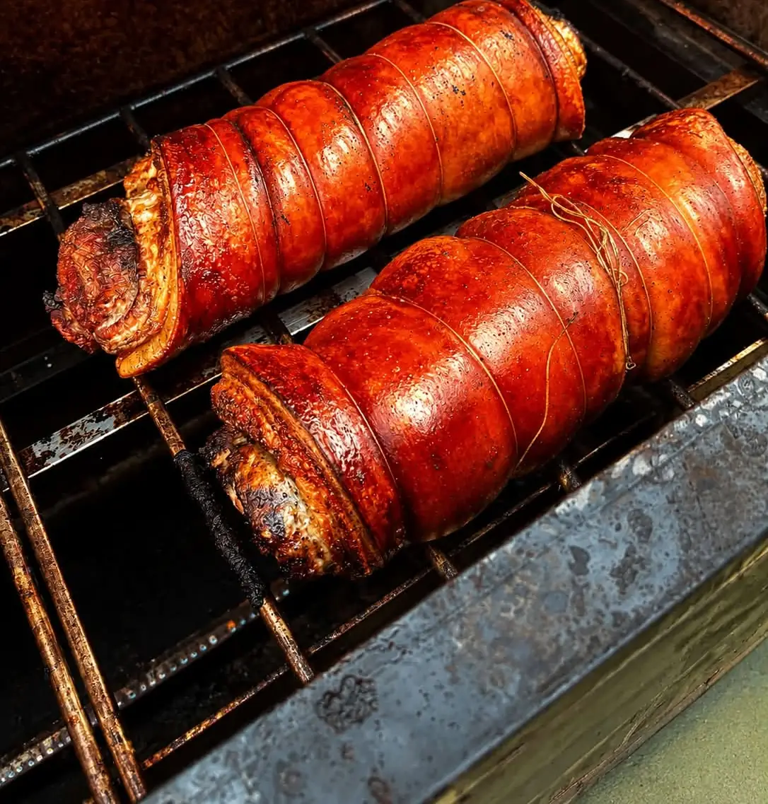 Two lechon belly rolls turning over the coals, skin glossy and blistered