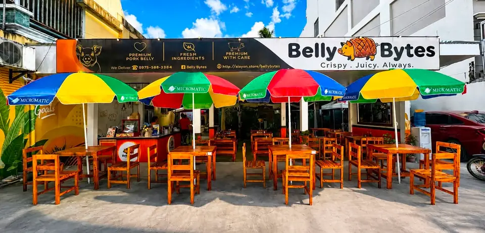 The front of Belly Bytes in daylight: the sign over the entrance, wooden tables and four bright umbrellas